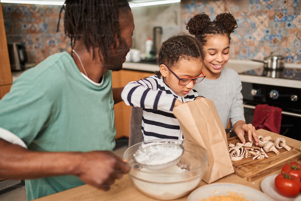 Happy young multi ethnic family with little daughters having fun while cooking at home together. Smiling parent and small multiracial children preparing dough for the pizza