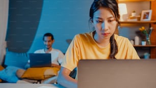 Freelance asia couple man and woman in casual wife focus financial work business in laptop computer on desk at night happy husband video call with family rest lay down after work on couch at home.