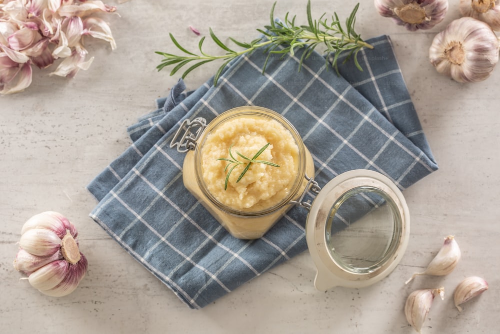 Draufsicht aromatische Knoblauchpaste in einem Glasgefäß auf rustikalem Küchentuch mit Zwiebeln und geschälten Nelken und Rosmarin.