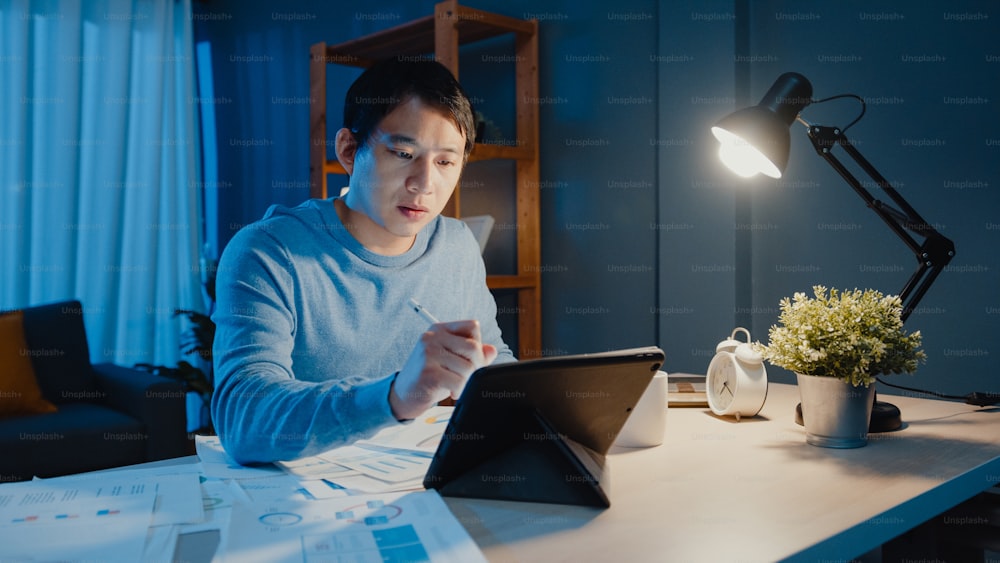 Empresário freelancer da Ásia foca trabalho caneta escrever em tablet ocupado com cheio de papelada gráfica na mesa na sala de estar em casa horas extras à noite, Trabalhar em casa durante o conceito de pandemia de COVID-19.