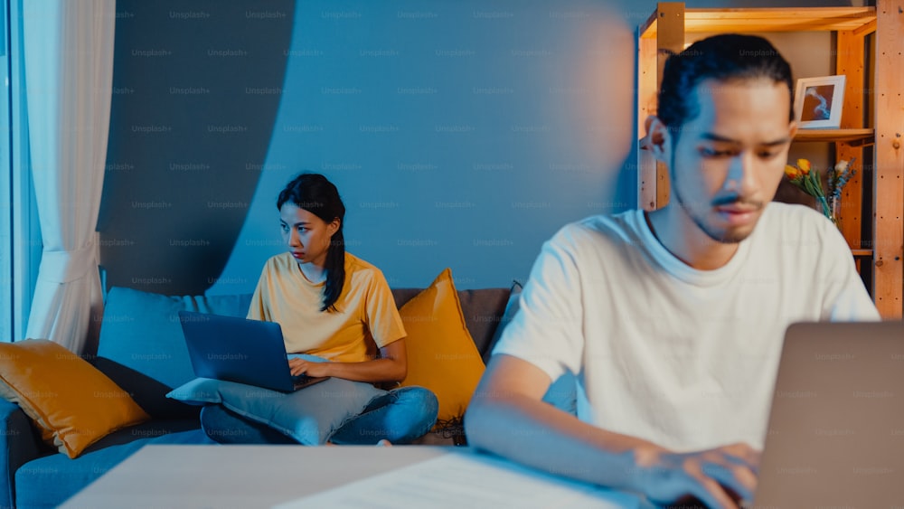 Freelance asia couple man and woman in casual feel serious focus on work separate laptop computer at night, hasband sit in front on table wife sit behind on couch at house. Work from home concept.