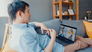 Joven empresario asiático que usa una computadora portátil habla con colegas sobre el plan en una reunión de videollamada mientras trabaja desde casa en la sala de estar. Autoaislamiento, distanciamiento social, cuarentena para la prevención del coronavirus.