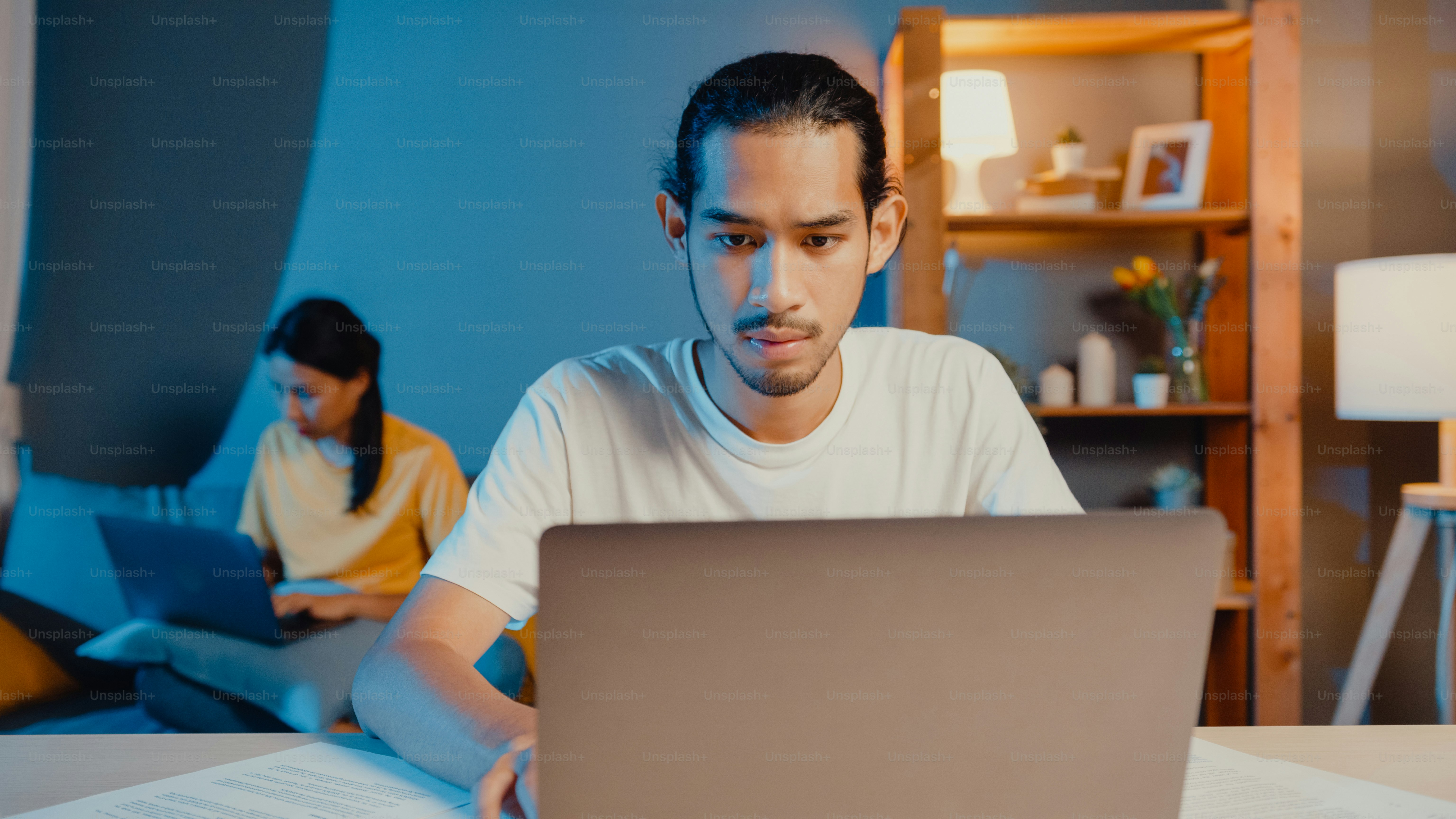 Pasangan freelance asia pria dan wanita dalam kasual merasa serius fokus pada pekerjaan komputer laptop terpisah di malam hari, hasband duduk di depan meja istri duduk di belakang di sofa di rumah. Konsep bekerja dari rumah.