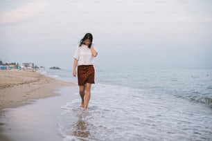 Femme hipster insouciante marchant pieds nus sur une plage de sable avec les vagues de la mer le soir, profitant d’un moment tranquille. Jeune femme décontractée se relaxant au bord de la mer à la station. Consciencieux