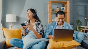 Happy young asian couple activity man use laptop computer work relax enjoy with women wear headphones use smartphone listen music at sofa in living room at house. Young married work from home concept.