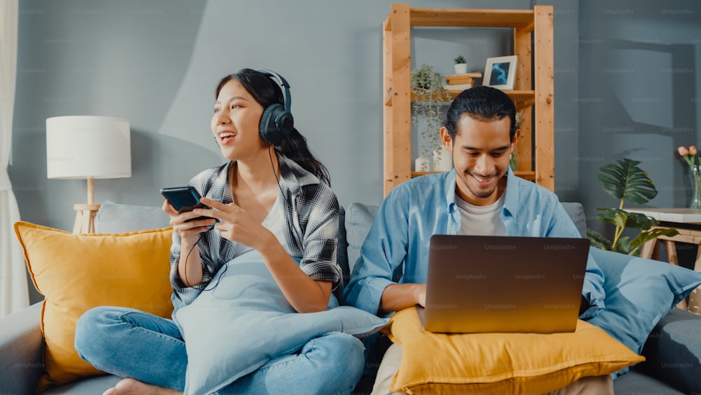 Happy young asian couple activity man use laptop computer work relax enjoy with women wear headphones use smartphone listen music at sofa in living room at house. Young married work from home concept.