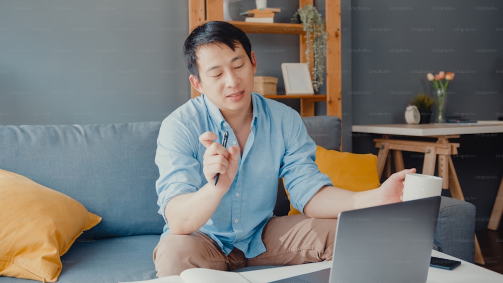 Giovane uomo d'affari asiatico che utilizza il laptop parla con i colleghi del piano in videochiamata mentre lavora in smart working da casa in salotto. Autoisolamento, distanziamento sociale, quarantena per la prevenzione del coronavirus.