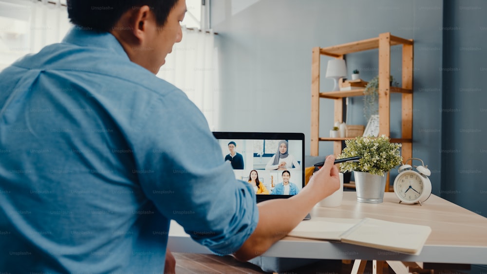 Joven empresario asiático que usa una computadora portátil habla con colegas sobre el plan en una reunión de videollamada mientras trabaja desde casa en la sala de estar. Autoaislamiento, distanciamiento social, cuarentena para la prevención del coronavirus.