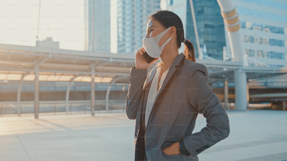 Young Asia businesswoman in fashion office clothe wear medical face mask talk via phone while walk alone outdoor in urban city. Business on go, Social distancing to prevent spread of COVID-19 concept.