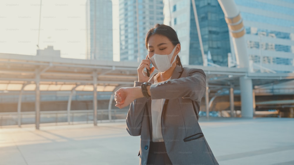 패션 사무실의 젊은 아시아 사업가가 도시 도시에서 혼자 야외를 걷는 동안 전화를 통해 의료 얼굴 마스크 이야기를 착용합니다. 이동 중 비즈니스, COVID-19 개념의 확산을 방지하기 위한 사회적 거리두기.