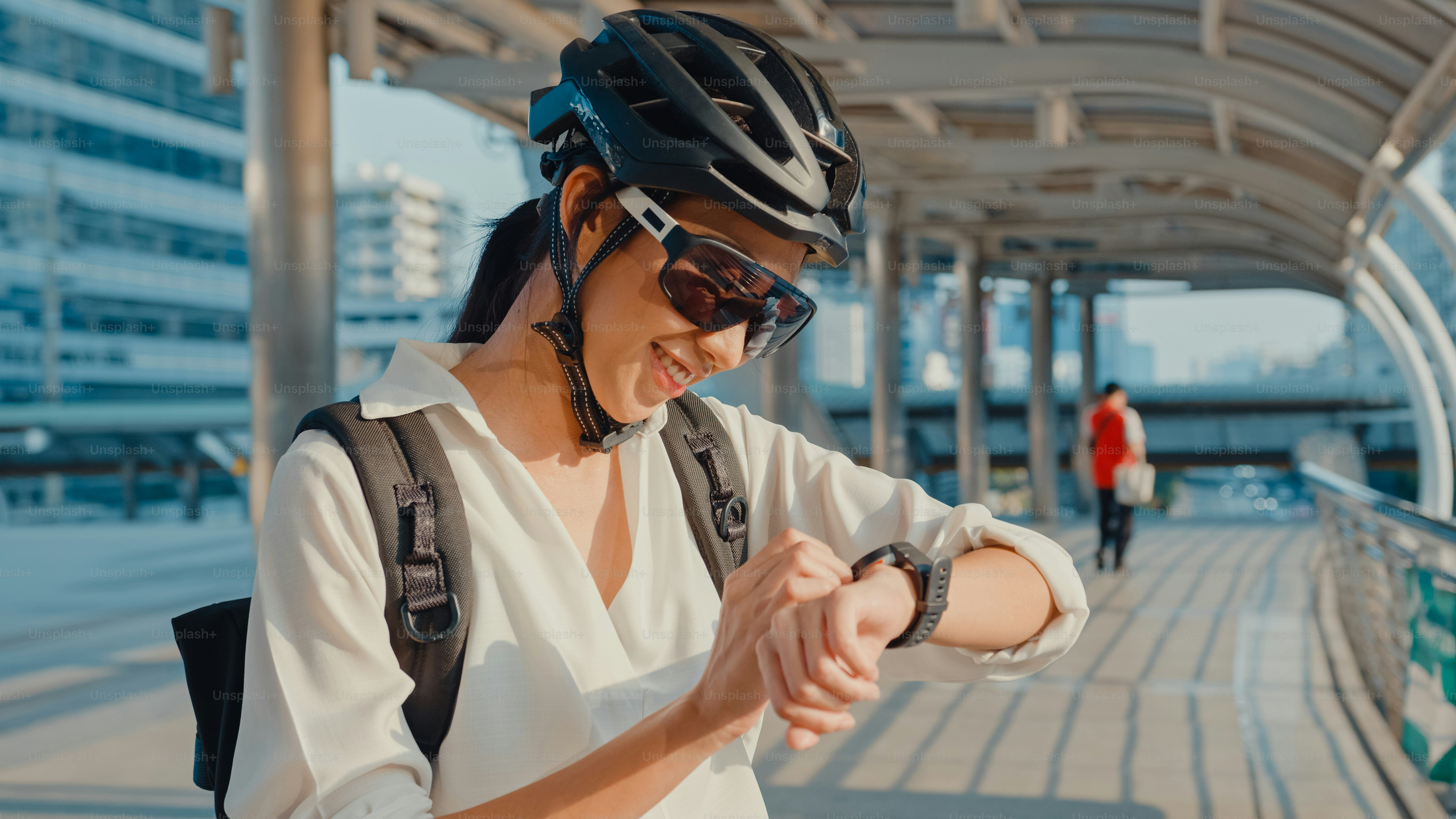 Asiatische Geschäftsfrau mit Rucksack Fahrrad lächeln Look Smartwatch in der Stadt Straße gehen zur Arbeit im Büro. Sportmädchen nutzen ihre Uhren-App für Fitness-Tracking. Pendeln zur Arbeit, Geschäftspendler in der Stadt.