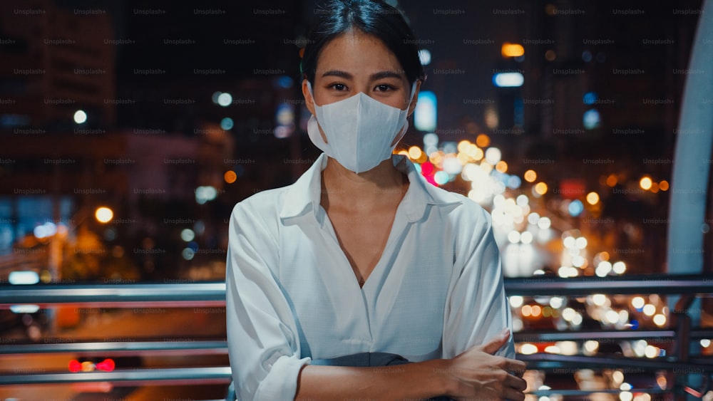 Successful young Asia businesswoman in fashion office clothes wear medical face mask smiling and looking at camera while happy stand alone outdoors in urban modern city night. Business on go concept.