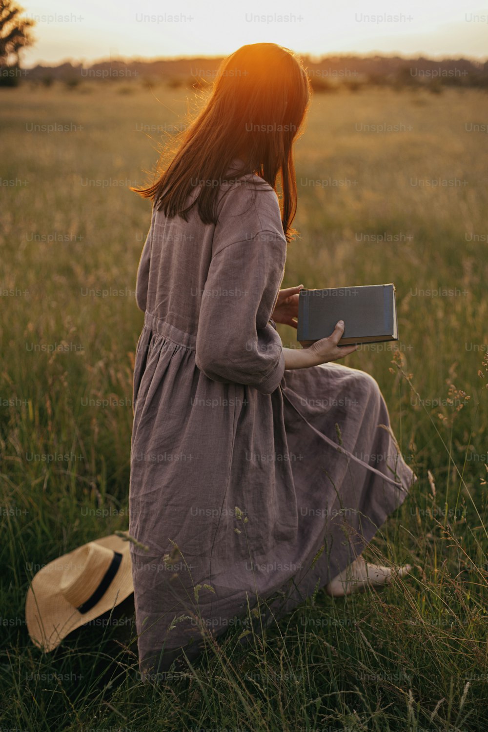 Bella donna in abito di lino che cammina con il libro in mano nel prato estivo al tramonto. Momento di calma atmosferica. Giovane femmina in vestito rustico che si rilassa nella campagna di sera