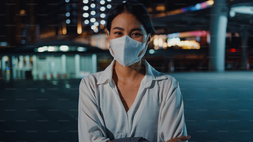 Successful young Asia businesswoman in fashion office clothes wear medical face mask smiling and looking at camera while happy stand alone outdoors in urban modern city night. Business on go concept.