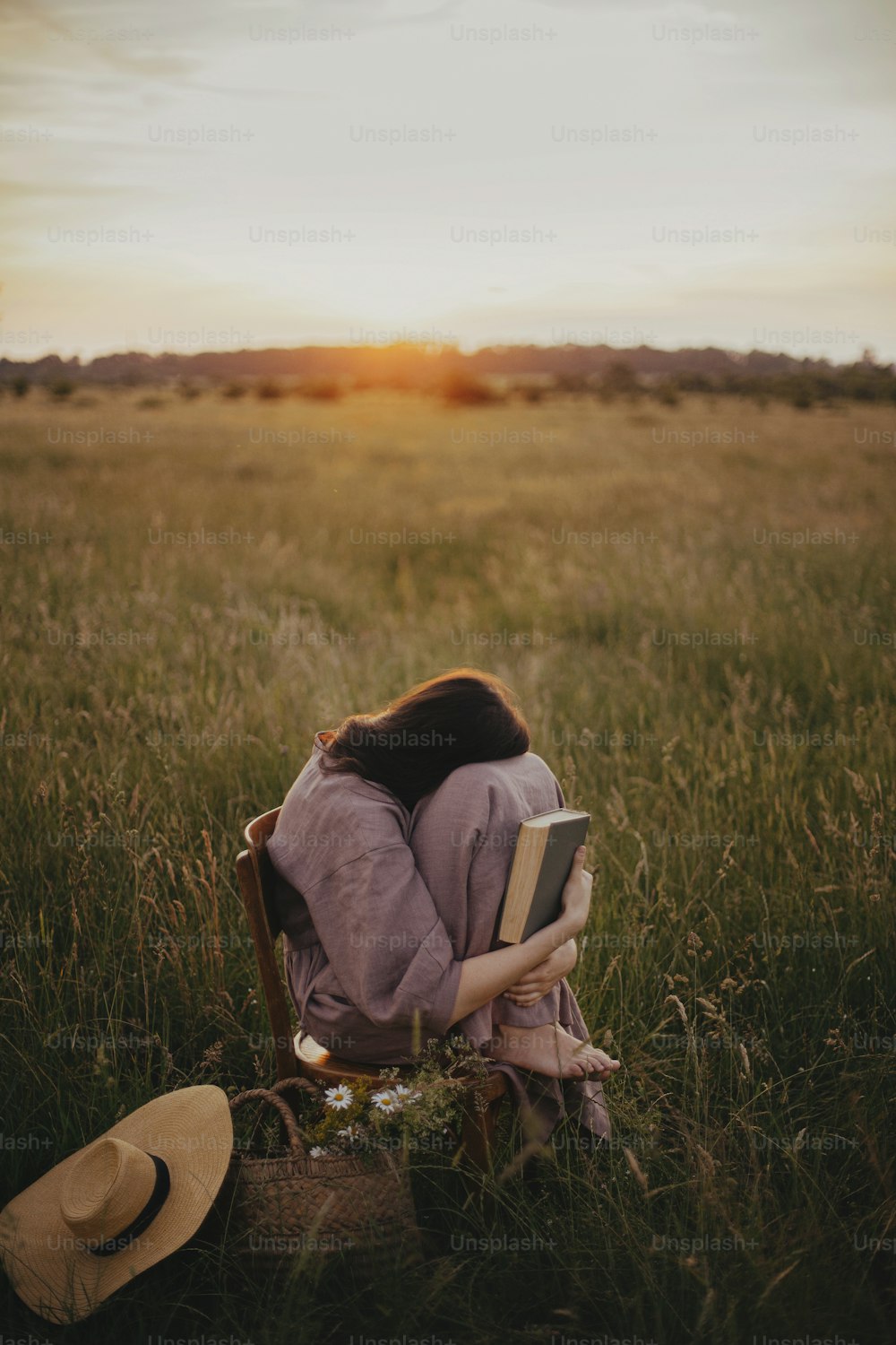 Beautiful woman in linen dress sitting on rustic chair and dreaming in summer meadow. Young female relaxing with book and basket of flowers in countryside sunset. Atmospheric tranquil moment