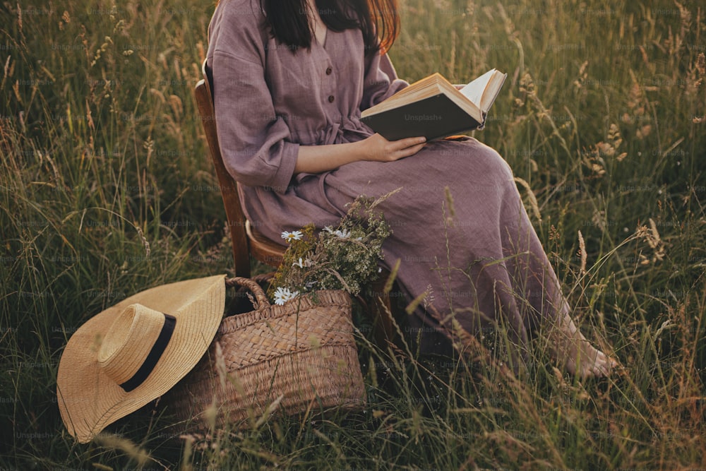 Beautiful woman in linen dress with book and basket of flowers sitting on wooden chair in summer meadow, close up. Young female relaxing in evening countryside. Atmospheric calm moment