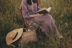 Beautiful woman in linen dress with book and basket of flowers sitting on wooden chair in summer meadow, close up. Young female relaxing in evening countryside. Atmospheric calm moment