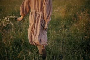 Beautiful woman in linen dress walking with wildflowers in hand in summer meadow in sunset, back view. Atmospheric calm moment. Young female in rustic dress relaxing in evening in countryside