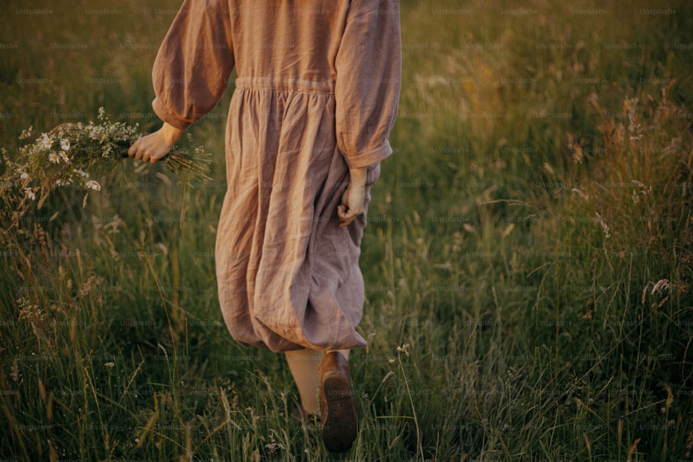 Bella donna in abito di lino che cammina con fiori selvatici in mano nel prato estivo al tramonto, vista posteriore. Momento di calma atmosferica. Giovane femmina in vestito rustico che si rilassa di sera in campagna