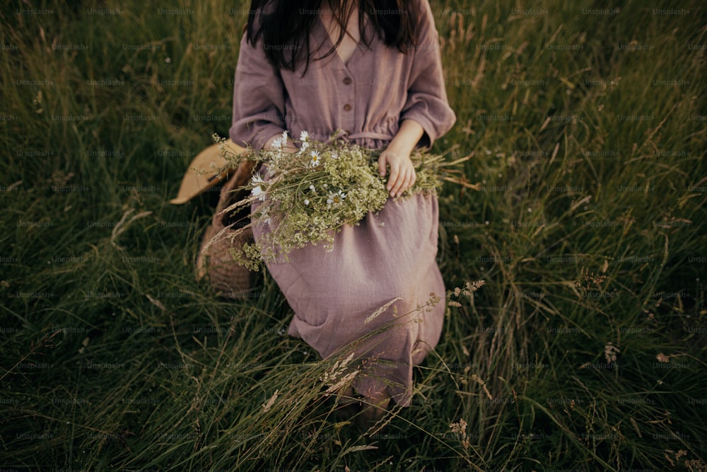 Beautiful woman with bouquet of wildflowers sitting barefoot on rustic chair among grass in summer meadow. Young female in linen dress relaxing in countryside. Atmospheric moment