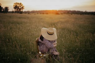 Bella donna in abito di lino e cappello di paglia seduta su sedia rustica nel prato estivo. Giovane femmina che si rilassa con cesto di fiori al tramonto in campagna. Momento di tranquillità atmosferica