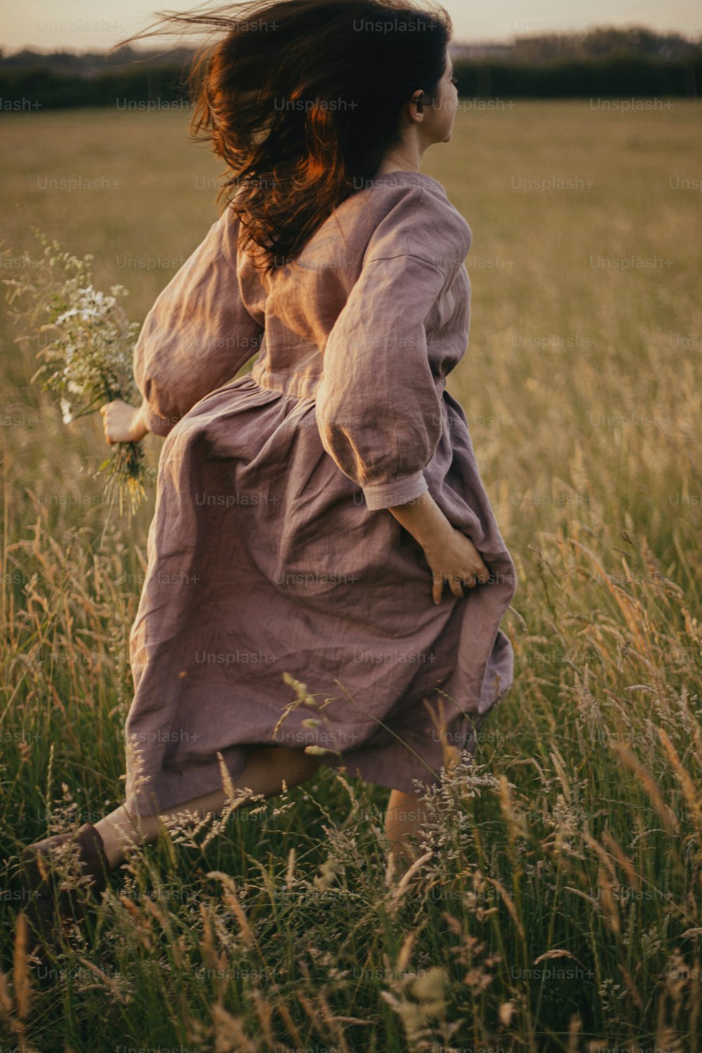 Bella donna in abito di lino che corre con fiori di campo in mano nel prato estivo al tramonto. Momento atmosferico spensierato. Giovane donna elegante in abito rustico che si gode una serata libera in campagna