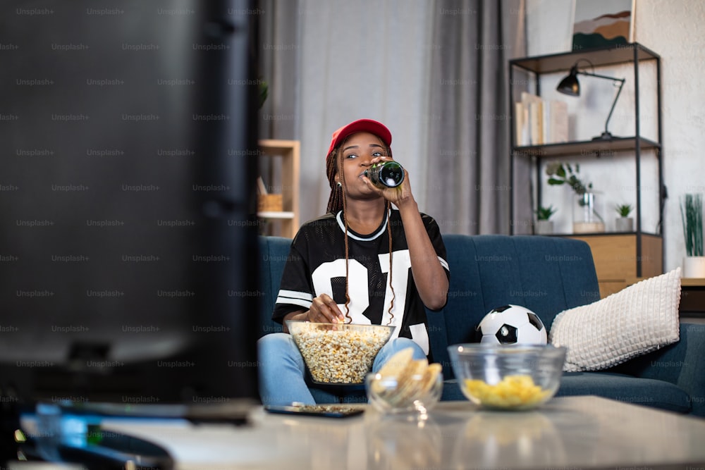 Mujer afroamericana con gorra roja bebiendo cerveza y comiendo bocadillos mientras ve un partido de fútbol en la televisión. Concepto de entretenimiento, aficionado y deporte.