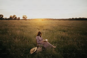 Beautiful woman in linen dress reading book in summer meadow in sunset. Young female with book and basket of flowers sitting on rustic chair and relaxing in countryside. Atmospheric calm moment
