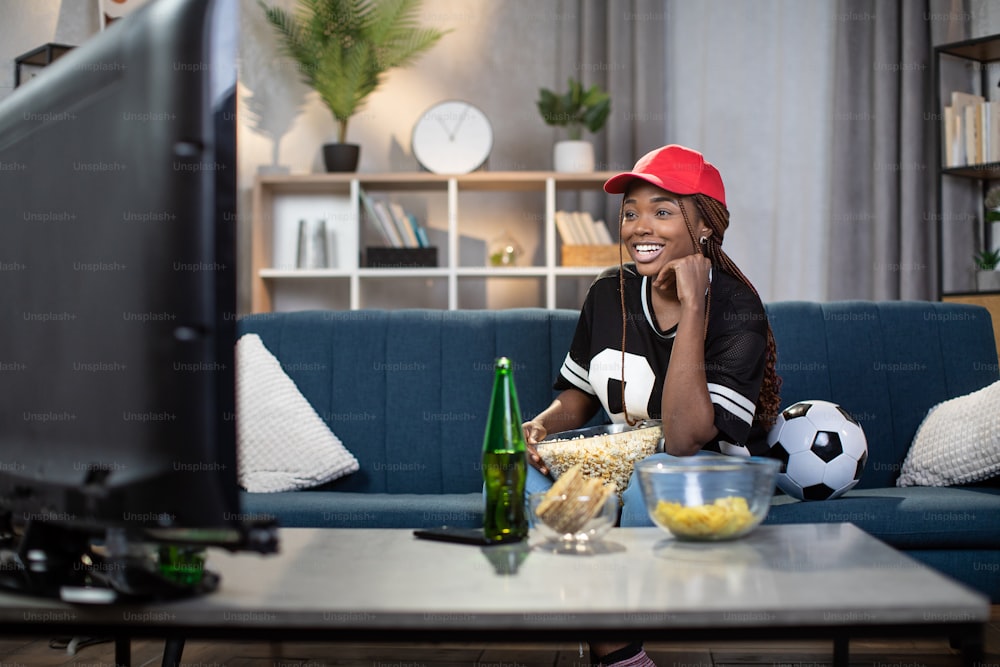 Happy african american woman in red cap sitting with beer and snacks on couch and watching football match. Young female spending free time alone with fun.