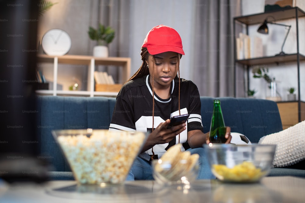 Mujer afroamericana que usa un teléfono inteligente moderno para ver la transmisión en vivo del campeonato europeo de fútbol mientras se queda en casa. Concepto de personas, tecnología y deporte.