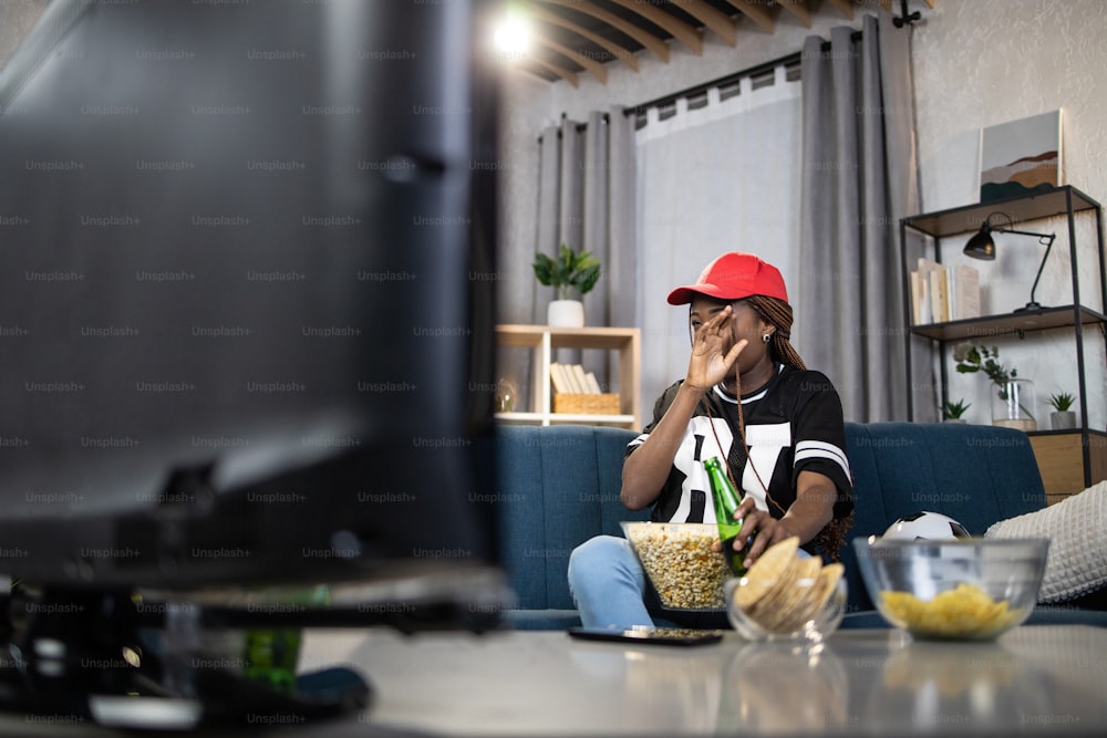 Donna attraente che guarda da parte e copre il viso con il palmo della mano durante il momento emotivo della partita di calcio. Femmina africana seduta a casa e guardando la TV.