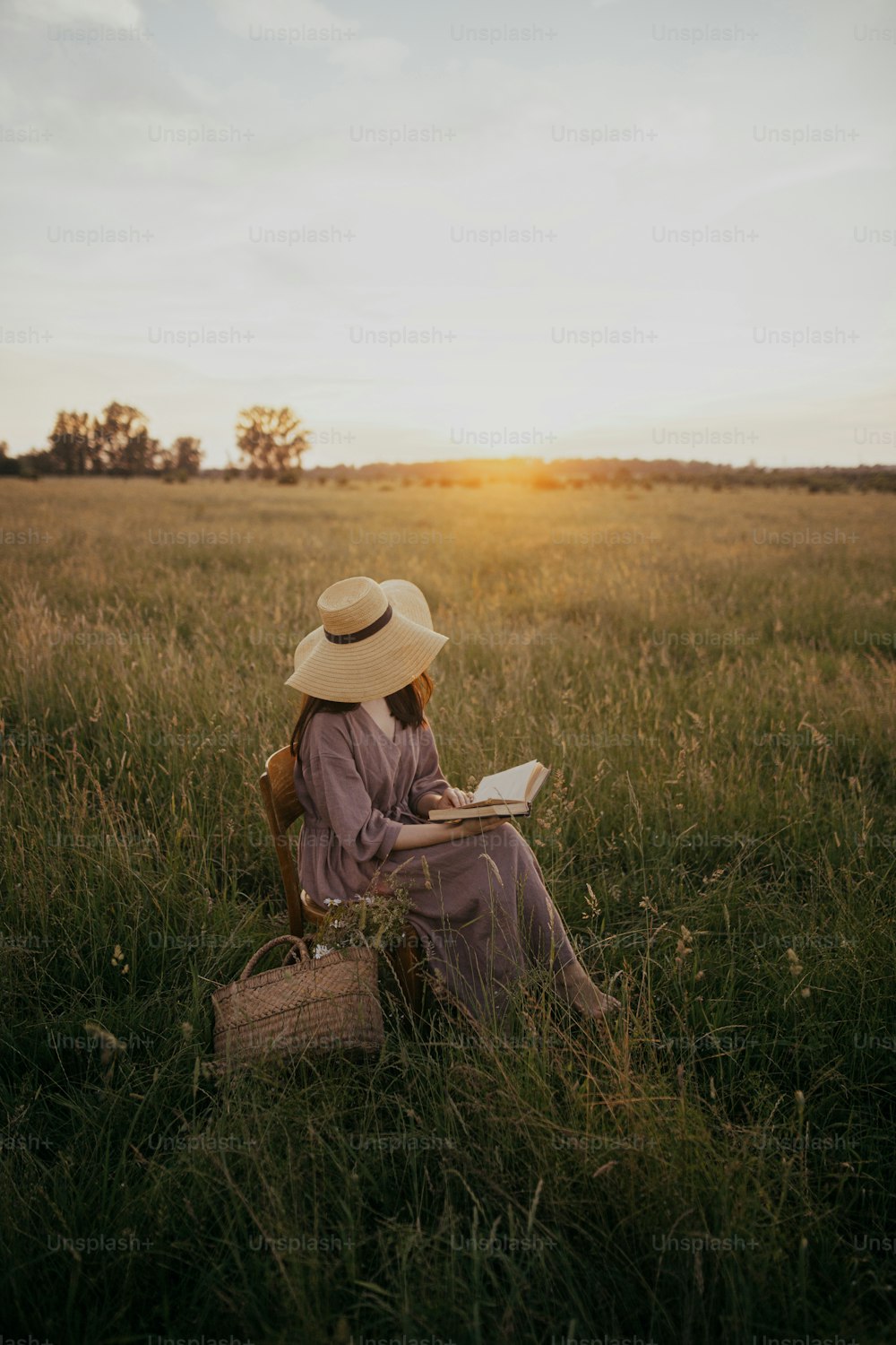 Beautiful woman in hat and linen dress with book and basket of flowers sitting on rustic chair in summer meadow in sunset. Atmospheric calm moment. Young female reading book in countryside