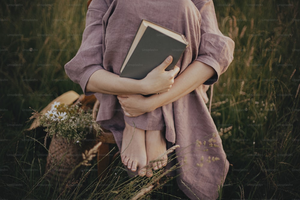Beautiful woman in linen dress sitting barefoot on rustic chair with book in summer meadow, close up. Young female holding book and relaxing in countryside. Atmospheric calm moment