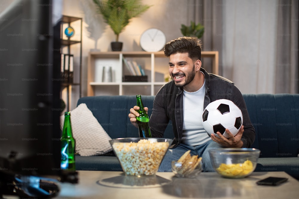 Aufgeregter indischer Mann mit Pommes und Bier, der auf einem gemütlichen Sofa sitzt und Sportfernsehen schaut. Junger bärtiger Kerl, der die Lieblingsfußballmannschaft während der Weltmeisterschaft unterstützt.