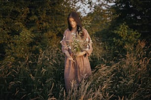 Mujer hermosa en vestido de lino recogiendo flores silvestres en el prado de verano en la tarde soleada. Momento rural atmosférico. Mujer joven con estilo en vestido rústico recogiendo flores en el campo. Vida lenta