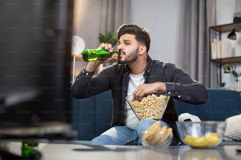 Cara indiano elegante bebendo cerveja gelada enquanto se senta confortavelmente no sofá e desfruta do campeonato de futebol em casa. Conceito de tempo livre, esporte e entretenimento.