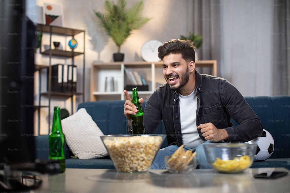 Hombre indio emocionado con patatas fritas y cerveza sentado en un cómodo sofá y viendo la televisión deportiva. Joven barbudo apoyando al equipo de fútbol favorito durante la copa del mundo.