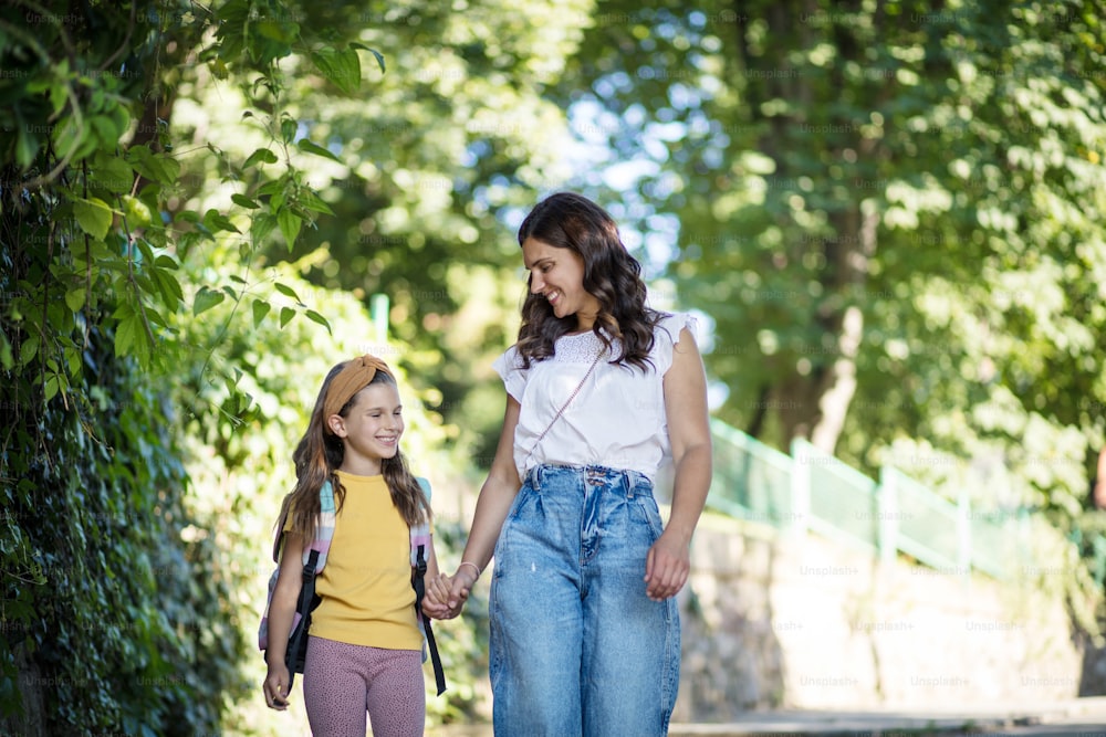 어린 소녀와 그녀의 어머니는 손을 잡고 자연을 걷고 있습니다. 학교 가방을 들고 다니는 어린 소녀.