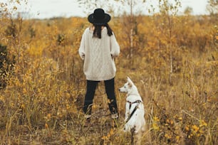 秋の木の背景にかわいい白い犬と一緒に旅行するスタイリッシュなヒップスターの女性。放浪癖。セーターと帽子をかぶった美しい若い女性がスイス羊飼いの子犬と一緒に立っています。背面図、コピースペース