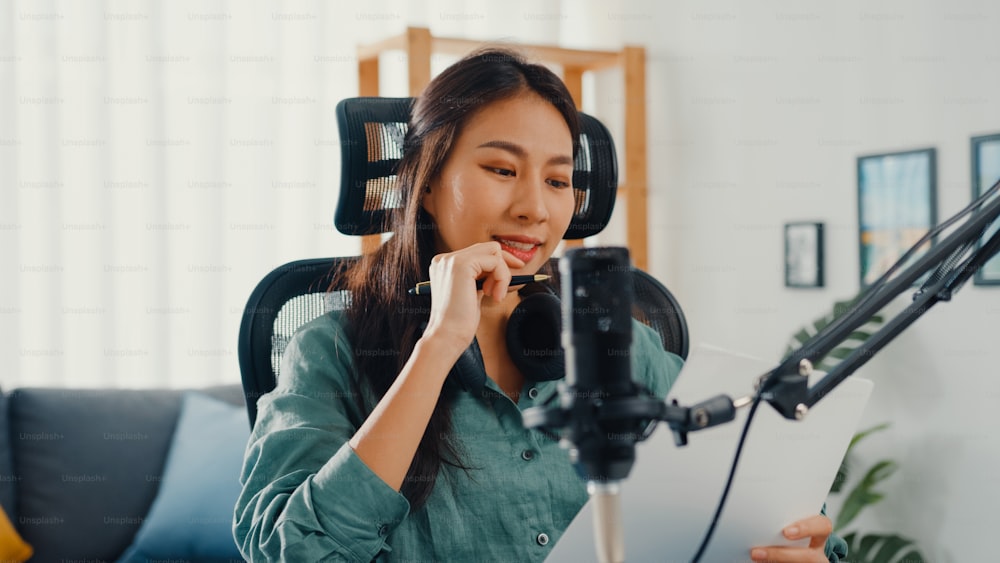 Attraente ragazza asiatica registra podcast usa il microfono tenere la carta creando contenuti per audio blog talk e pratica per l'argomento di revisione nella sua stanza. Crea podcast audio da casa, concetto di attrezzatura audio.