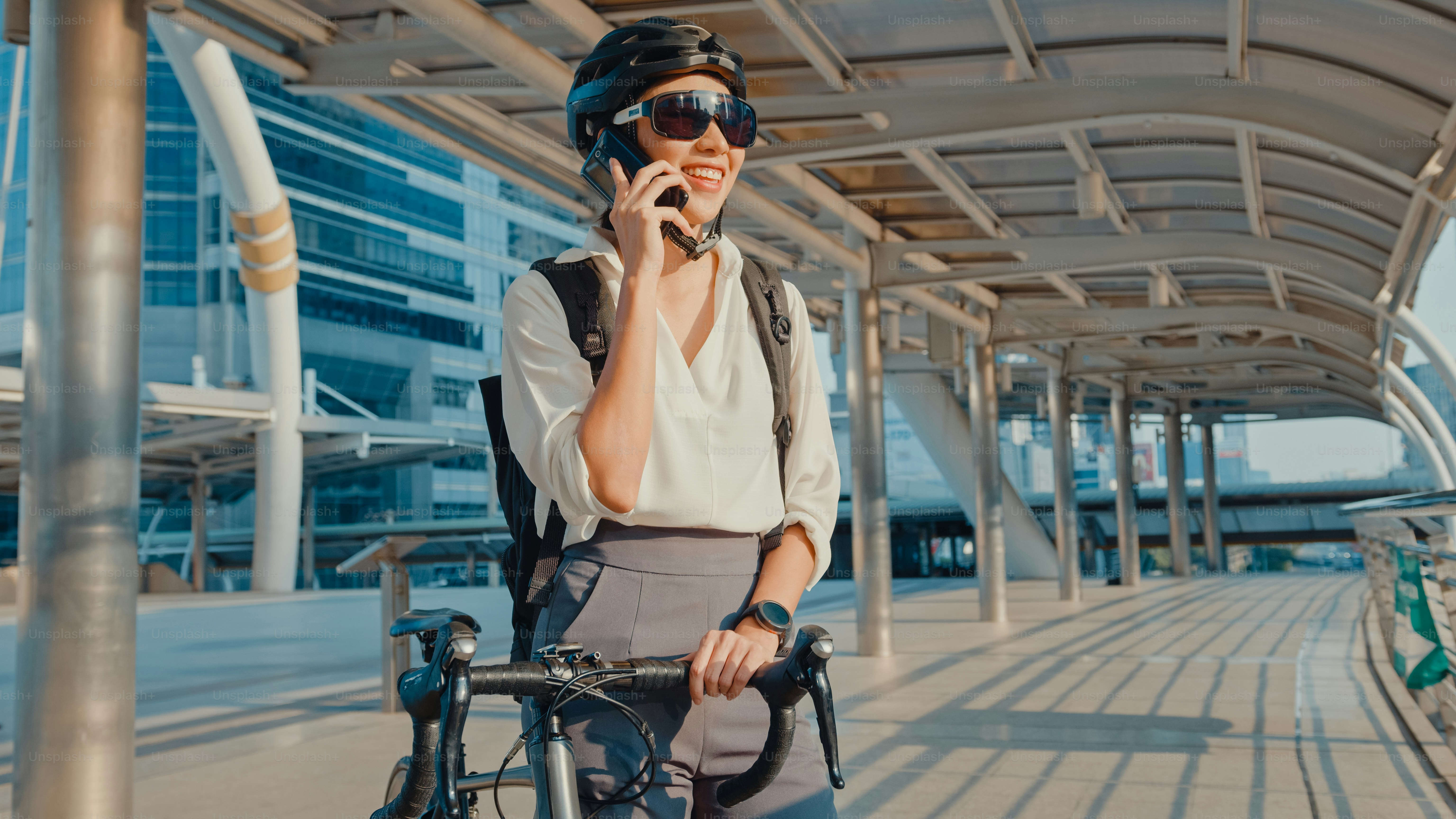 Asiatische Geschäftsfrau mit Rucksack Anruf Handy sprechen lächelnd in der Stadt Straße gehen zur Arbeit im Büro. Sportmädchen benutzen ihr Telefon für die Arbeit im Geschäft. Mit dem Fahrrad zur Arbeit pendeln, Business-Pendler in der Stadt.
