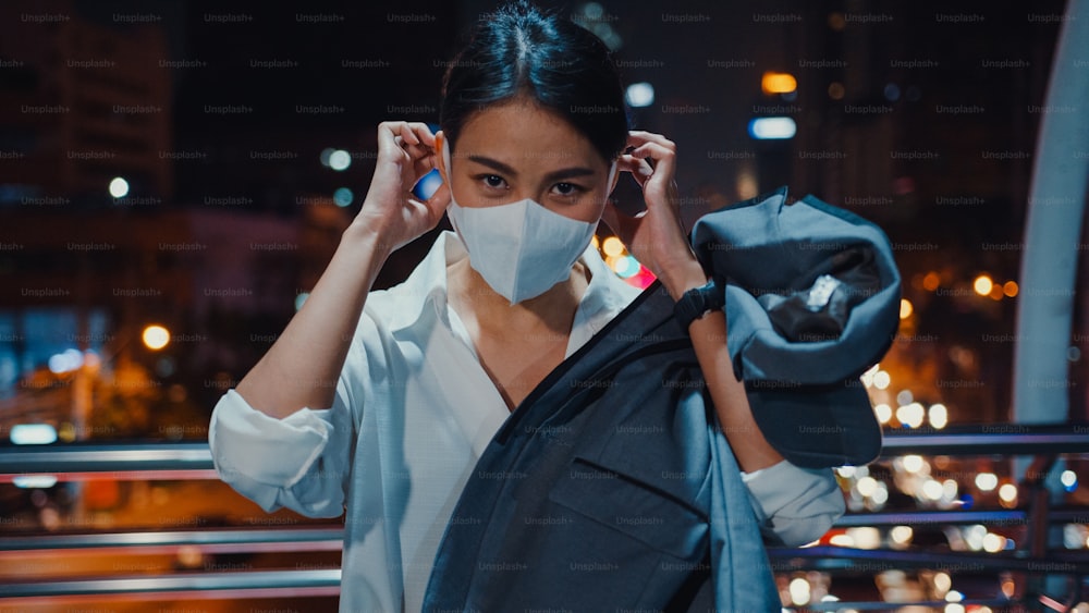 Successful young Asia businesswoman in fashion office clothes wear medical face mask smiling and looking at camera while happy stand alone outdoors in urban modern city night. Business on go concept.