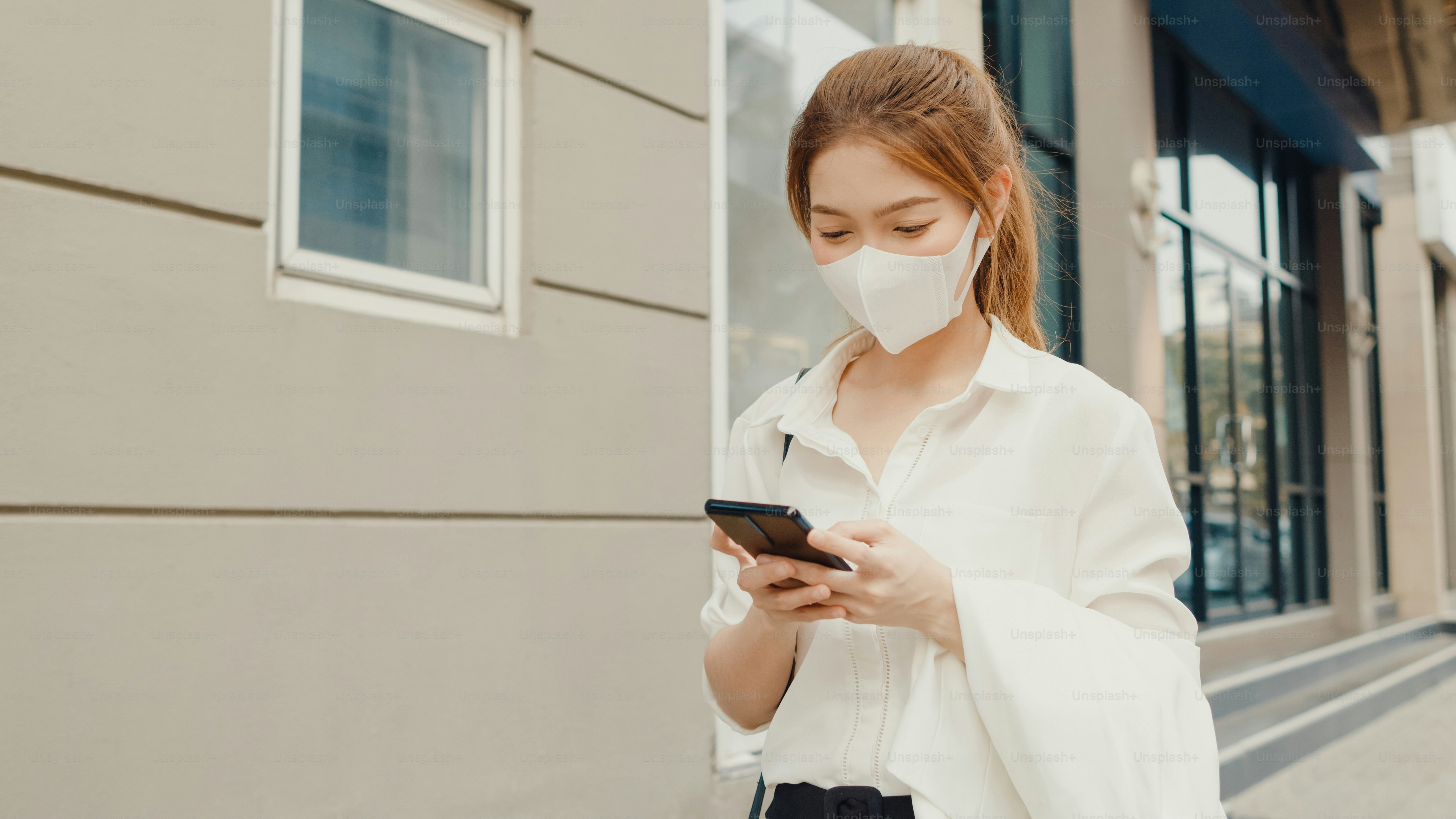Pengusaha muda Asia yang sukses dengan pakaian kantor fashion mengenakan masker wajah medis menggunakan ponsel pintar sambil berjalan sendirian di luar ruangan di kota modern perkotaan di pagi hari. Konsep bisnis saat bepergian.