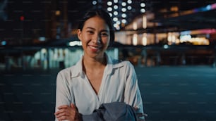 Successful young Asia businesswoman in fashion office clothes smiling and looking at camera while happy standing alone outdoors in urban modern city at night. Business on the go and commuter concept.