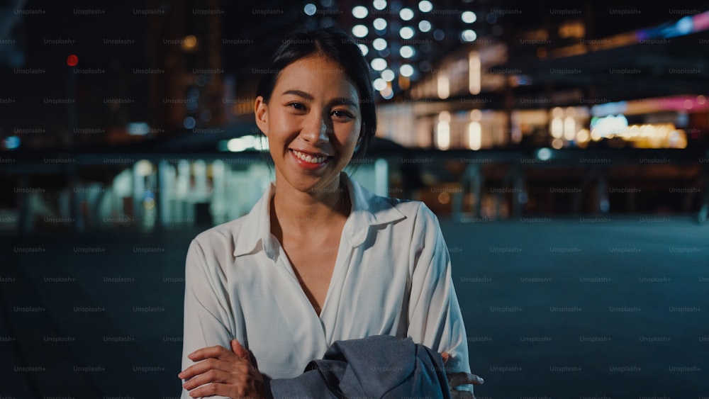 Successful young Asia businesswoman in fashion office clothes smiling and looking at camera while happy standing alone outdoors in urban modern city at night. Business on the go and commuter concept.