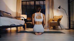 Young Asia lady in sportswear doing yoga exercise working out in living room at home at night. Sport and recreation activity, social distancing, quarantine for corona virus prevention concept.