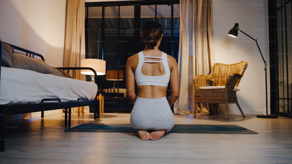 Jeune femme asiatique en vêtements de sport faisant de l’exercice de yoga dans le salon à la maison la nuit. Activité sportive et récréative, distanciation sociale, quarantaine pour le concept de prévention du virus corona.