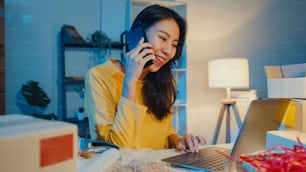 Young Asia woman call smartphone talk with customer for check confirm order in stock on laptop computer in home office at night. Small business, online market delivery, lifestyle freelance concept.