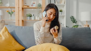 Sick young Asian woman hold medicine sit on couch use smartphone call to consult with doctor at home. Girl take medicine after doctor order, quarantine at home, Social distancing coronavirus concept.