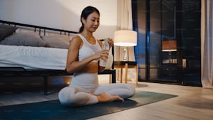 Young Asia lady doing yoga exercise working out and drinking pure water in living room at home night. Sport and recreation activity, social distancing, quarantine for corona virus prevention concept.